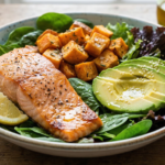 Seared salmon fillet seasoned with black pepper served over a bed of fresh spinach and mixed greens, topped with roasted sweet potato cubes, sliced avocado, and lemon wedges, drizzled with olive oil in a speckled ceramic bowl on a rustic wooden table.